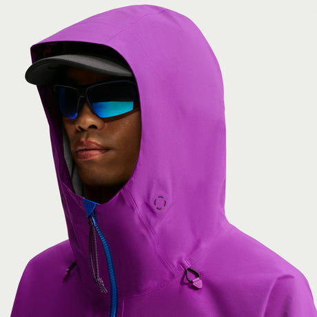 A close-up of a person wearing a bright purple hooded jacket, blue mirrored sunglasses, and a black cap against a light gray background. - Nike ACG Misery Ridge Storm-FIT ADV GORE-TEX Jacket (Men's) - Bold Berry/Hyper Royal/Lemon Venom