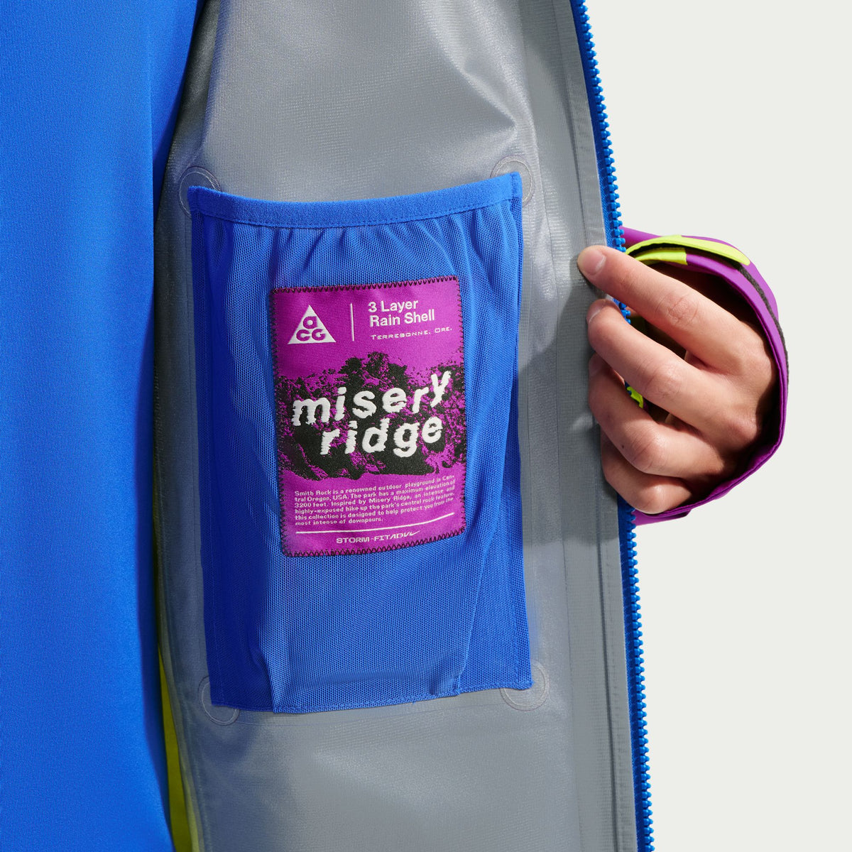 Close-up of a gray waterproof jacket with a bright blue inner pocket and a pink-and-purple Misery Ridge 3-Layer Rain Shell patch, shown on a model whose hand is holding the zipper. - Nike ACG Misery Ridge Storm-FIT ADV GORE-TEX Jacket (Men's) - Bold Berry/Hyper Royal/Lemon Venom