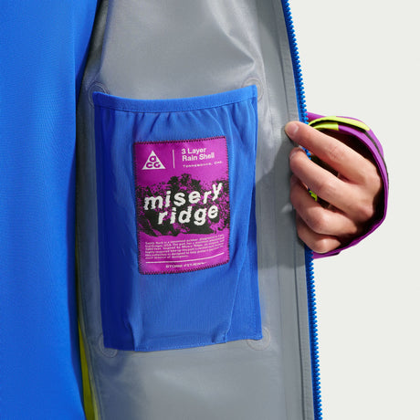 Close-up of a gray waterproof jacket with a bright blue inner pocket and a pink-and-purple Misery Ridge 3-Layer Rain Shell patch, shown on a model whose hand is holding the zipper. - Nike ACG Misery Ridge Storm-FIT ADV GORE-TEX Jacket (Men's) - Bold Berry/Hyper Royal/Lemon Venom