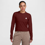 A woman modeling a long-sleeve crewneck shirt in deep burgundy with a small white geometric logo on the left chest, paired with beige pants, against a light gray studio background. - Nike ACG Dri-FIT Long-Sleeve Top (Women's) - Bold Berry/Summit White