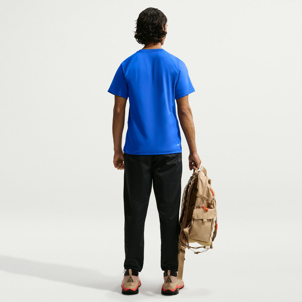 A man is shown from behind wearing a bright blue athletic t-shirt, black pants, and hiking shoes while carrying a beige backpack against a clean white studio background. - Nike ACG Dri-FIT Short-Sleeve Base Layer Top (Men's) - Hyper Royal/Summit White
