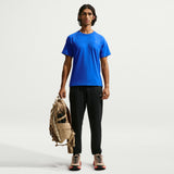 A man stands against a light gray studio background wearing a bright blue short-sleeve athletic T-shirt, black jogger pants, and running shoes, while holding a tan backpack. - Nike ACG Dri-FIT Short-Sleeve Base Layer Top (Men's) - Hyper Royal/Summit White