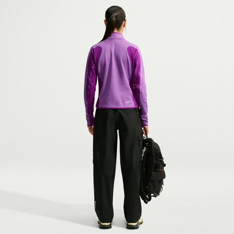 A woman shown from the back wearing a purple long-sleeve athletic jacket and black pants, holding a black backpack against a clean light gray studio background. - Nike ACG Wolf Grid Therma-FIT 1/2-Zip Trail Hiking Top (Women's) - Monarch Purple/Bold Berry/Summit White