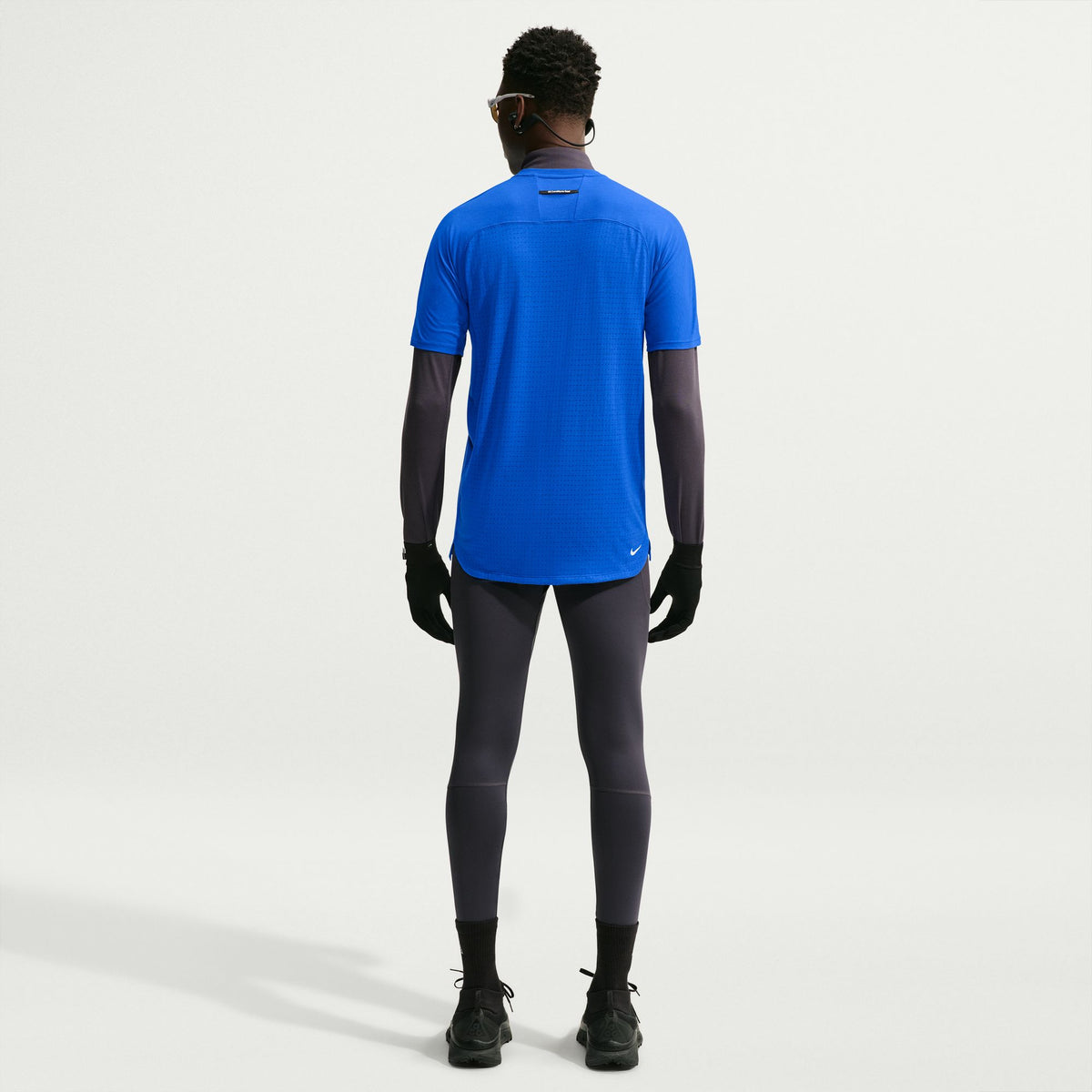 A man is shown from the back wearing a bright blue short-sleeve athletic shirt over a dark gray long-sleeve base layer, paired with dark gray leggings, black gloves, and black running shoes, against a plain light gray studio background. - Nike ACG Solar Chase Dri-FIT ADV Trail Running Top (Men's) - Hyper Royal/Hyper Royal/Summit White