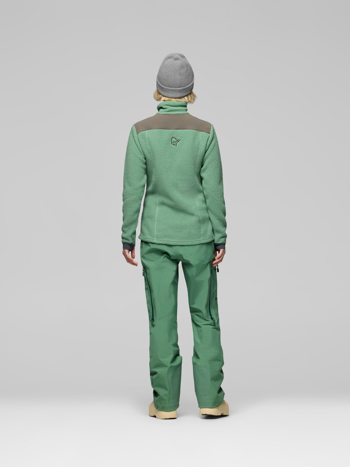 A person shown from the back wearing a green fleece jacket with brown shoulder panels and matching green pants, standing against a light gray studio background. - Norrøna Trollveggen Warm3 Jacket (Women's) - Dark Ivy