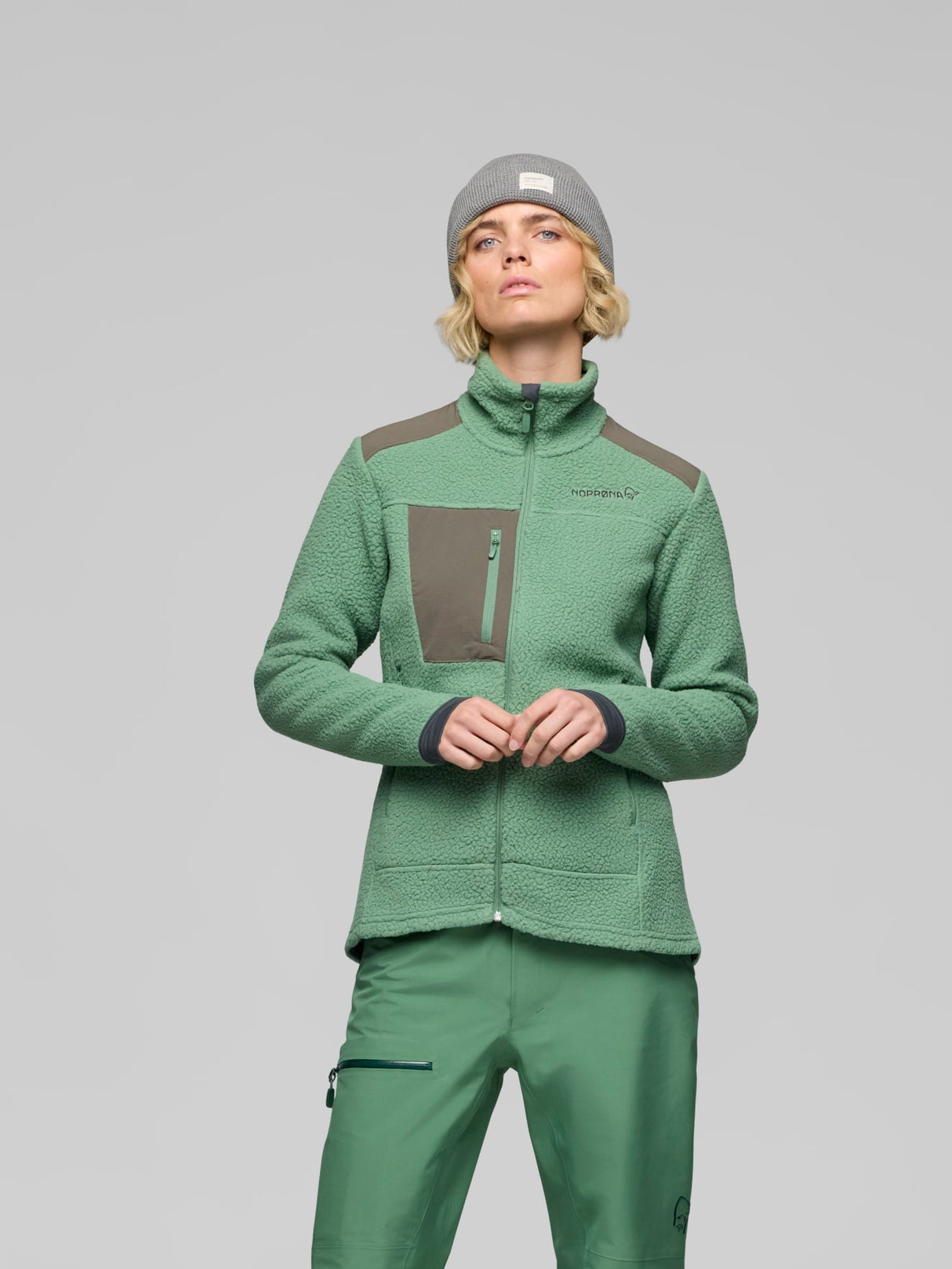 A model wearing a green fleece zip-up jacket with brown shoulder and chest panels, matching green pants, and a gray beanie, photographed against a solid gray studio background. - Norrøna Trollveggen Warm3 Jacket (Women's) - Dark Ivy