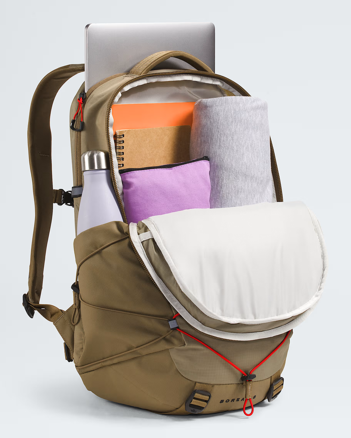 A tan BOREA backpack shown open, filled with travel items including a silver laptop, orange notebook, gray rolled blanket, purple pouch, and white bottle, on a light gray studio background. - The North Face Borealis Backpack - Cedar/Lava Red