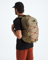 A man wearing a black cap, black t-shirt, and rust-orange shorts is shown from the back, wearing a beige The North Face backpack with red compression straps against a white studio background. - The North Face Borealis Backpack - Cedar/Lava Red