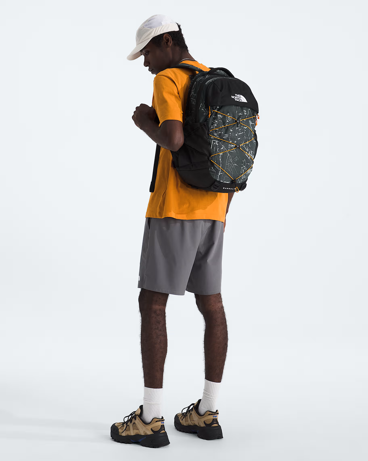 A man wearing a white cap, yellow t-shirt, gray shorts, and hiking shoes is shown from the back wearing a black and gray map-patterned backpack by The North Face, set against a plain white studio background. - The North Face Borealis Backpack - Anthracite Grey Conrad Topos Print/Summit Gold