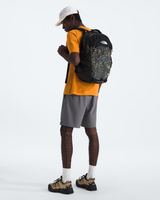 A man wearing a white cap, yellow t-shirt, gray shorts, and hiking shoes is shown from the back wearing a black and gray map-patterned backpack by The North Face, set against a plain white studio background. - The North Face Borealis Backpack - Anthracite Grey Conrad Topos Print/Summit Gold