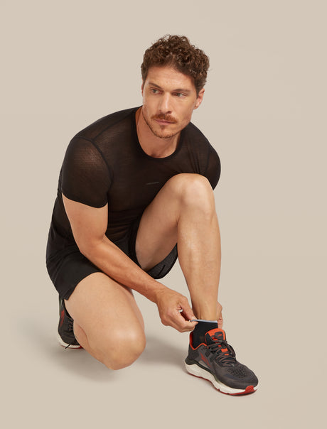 A man in a black athletic mesh t-shirt and black shorts crouches while tying the laces on a black and orange running shoe against a solid beige background. - Icebreaker Merino Multisport Light Mini Socks (Men's) - Black/Snow