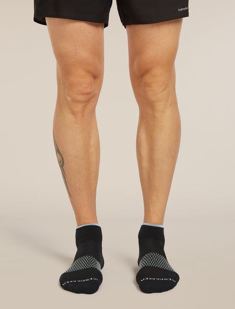 A person's legs wearing black athletic socks with gray textured grip panels on the toes and heels, paired with black athletic shorts, against a solid light beige background. - Icebreaker Merino Multisport Light Mini Socks (Men's) - Black/Snow