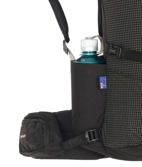 Close-up of a black backpack side pocket holding a dark blue water bottle with a white cap, showing the backpack's grid-patterned fabric and black straps against a white background. - One Planet Extrovert 55L Pack - Black