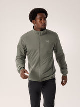 A man wearing a light olive green Arc'teryx zip-up jacket and black pants, standing against a plain light beige studio background. - Arc'teryx Atom Jacket (Men's) - Forage