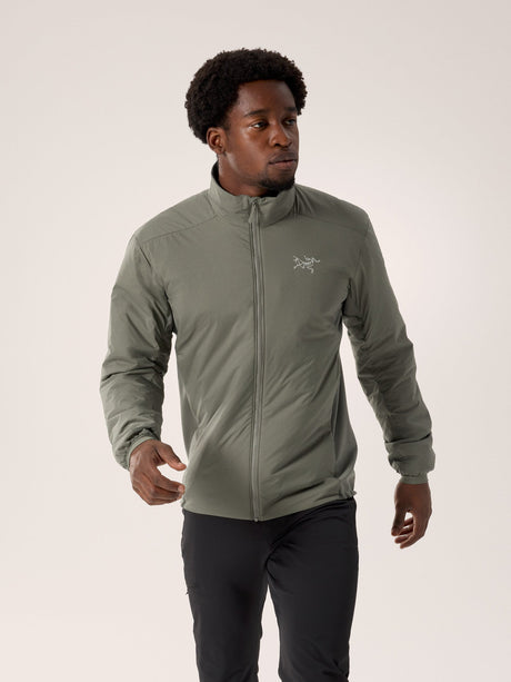 A man wearing a light olive green Arc'teryx zip-up jacket and black pants, standing against a plain light beige studio background. - Arc'teryx Atom Jacket (Men's) - Forage
