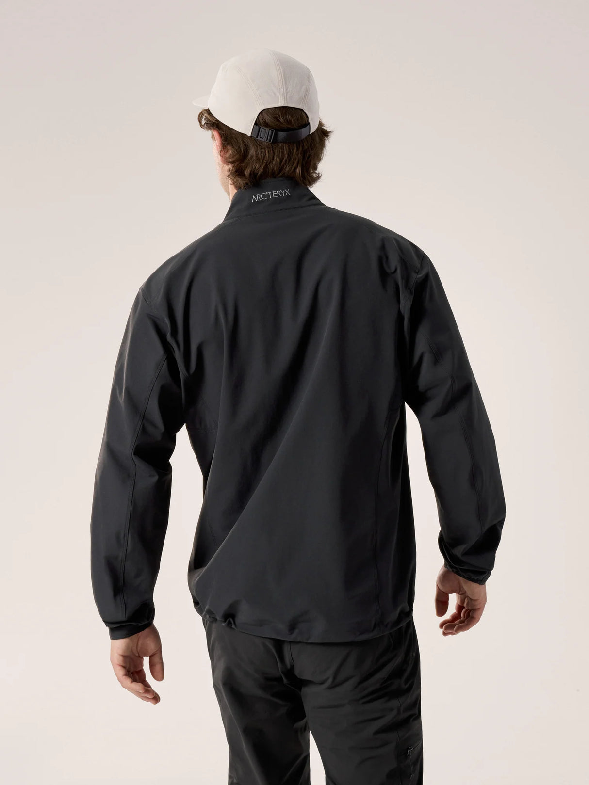 A man viewed from behind wearing a black Arc'teryx lightweight jacket, black pants, and a white baseball cap against a light beige studio background. - Arc'teryx Gamma Jacket (Men's) - Black