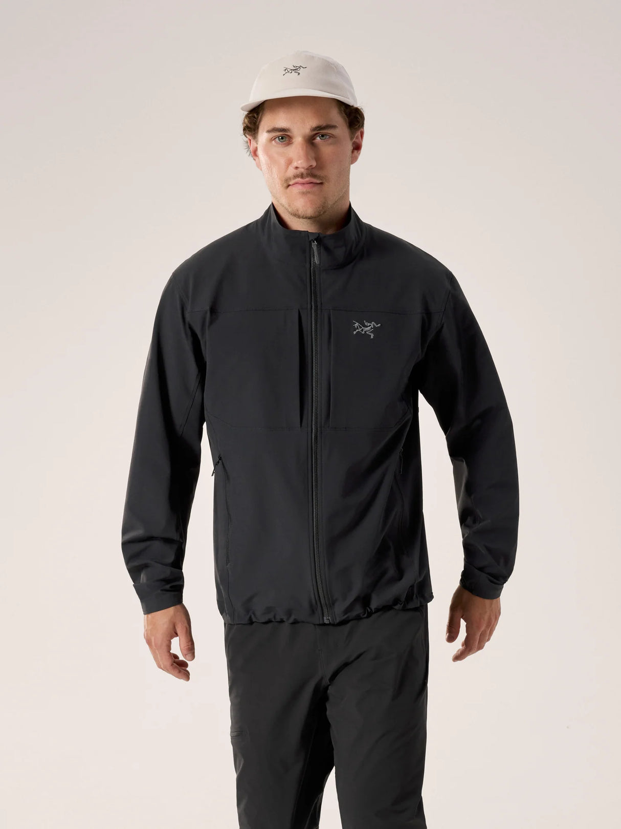 A man wearing a black Arc'teryx zip-up jacket and matching black pants, paired with a light beige baseball cap, standing against a plain light beige studio background. - Arc'teryx Gamma Jacket (Men's) - Black