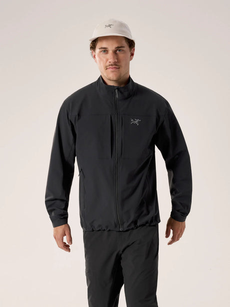 A man wearing a black Arc'teryx zip-up jacket and matching black pants, paired with a light beige baseball cap, standing against a plain light beige studio background. - Arc'teryx Gamma Jacket (Men's) - Black