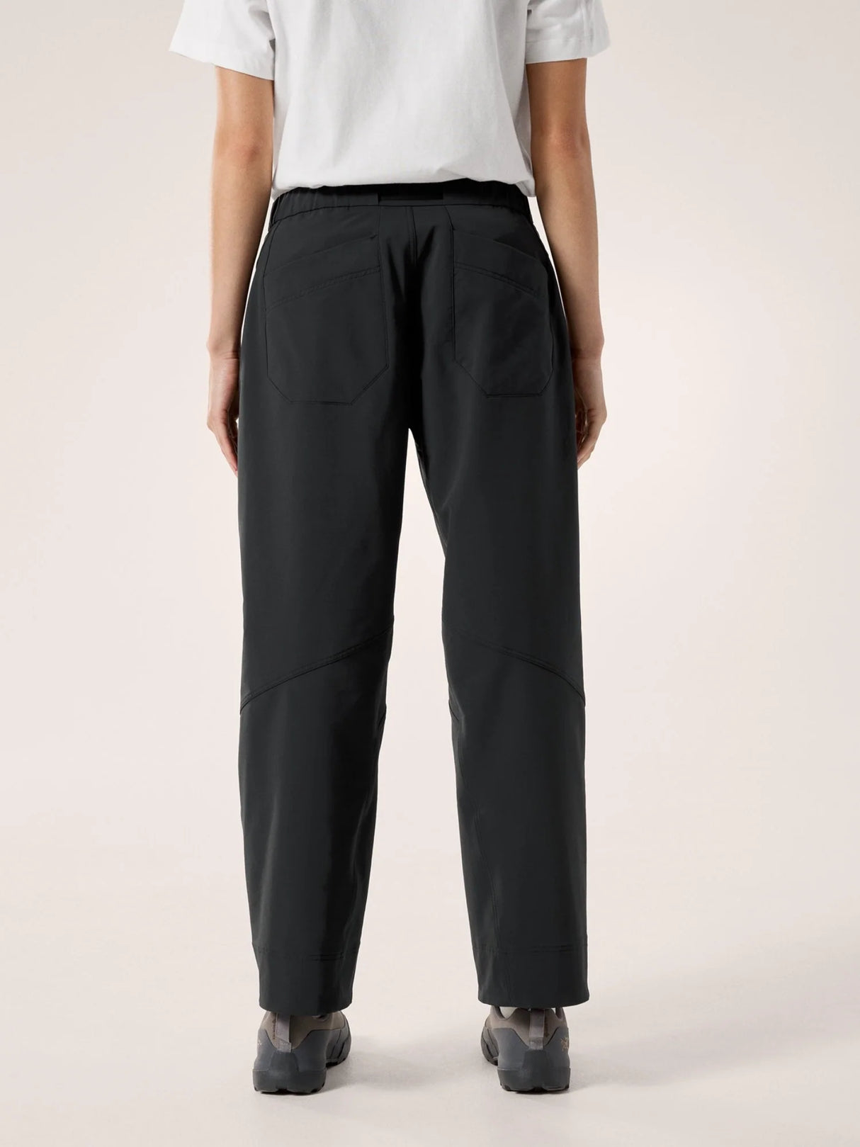 Rear view of a person wearing loose black trousers with a white short-sleeve top and dark shoes, photographed against a plain light beige studio background. - Arc'teryx Nia Pants (Women's) - Black