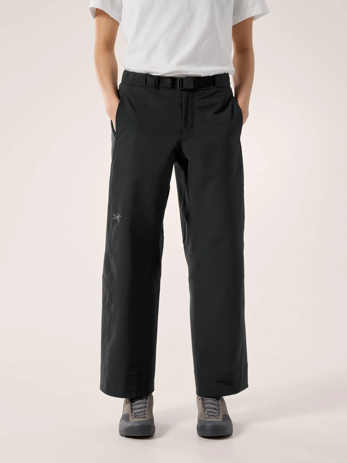 A person wearing black wide-leg trousers with a white t-shirt and gray sneakers, standing against a plain light beige studio background. - Arc'teryx Nia Pants (Women's) - Black