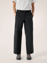 A person wearing black wide-leg trousers with a white t-shirt and gray sneakers, standing against a plain light beige studio background. - Arc'teryx Nia Pants (Women's) - Black
