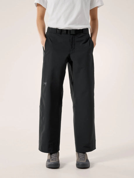A person wearing black wide-leg trousers with a white t-shirt and gray sneakers, standing against a plain light beige studio background. - Arc'teryx Nia Pants (Women's) - Black