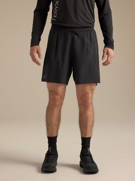 A model from the waist down wearing black athletic shorts, a black long-sleeve top, black socks, and black running shoes against a solid beige studio background. The shorts feature a drawstring waist and a small white logo on the left leg. - Arc'teryx Norvan Liner Shorts 7" (Men's) - Black