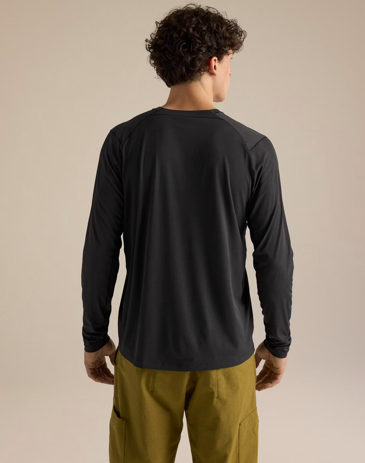 A man viewed from behind wearing a black long-sleeve athletic shirt and olive green cargo pants, standing against a plain beige studio background. - Arc'teryx Cormac Crew Neck Shirt Long Sleeve (Men's) - Black
