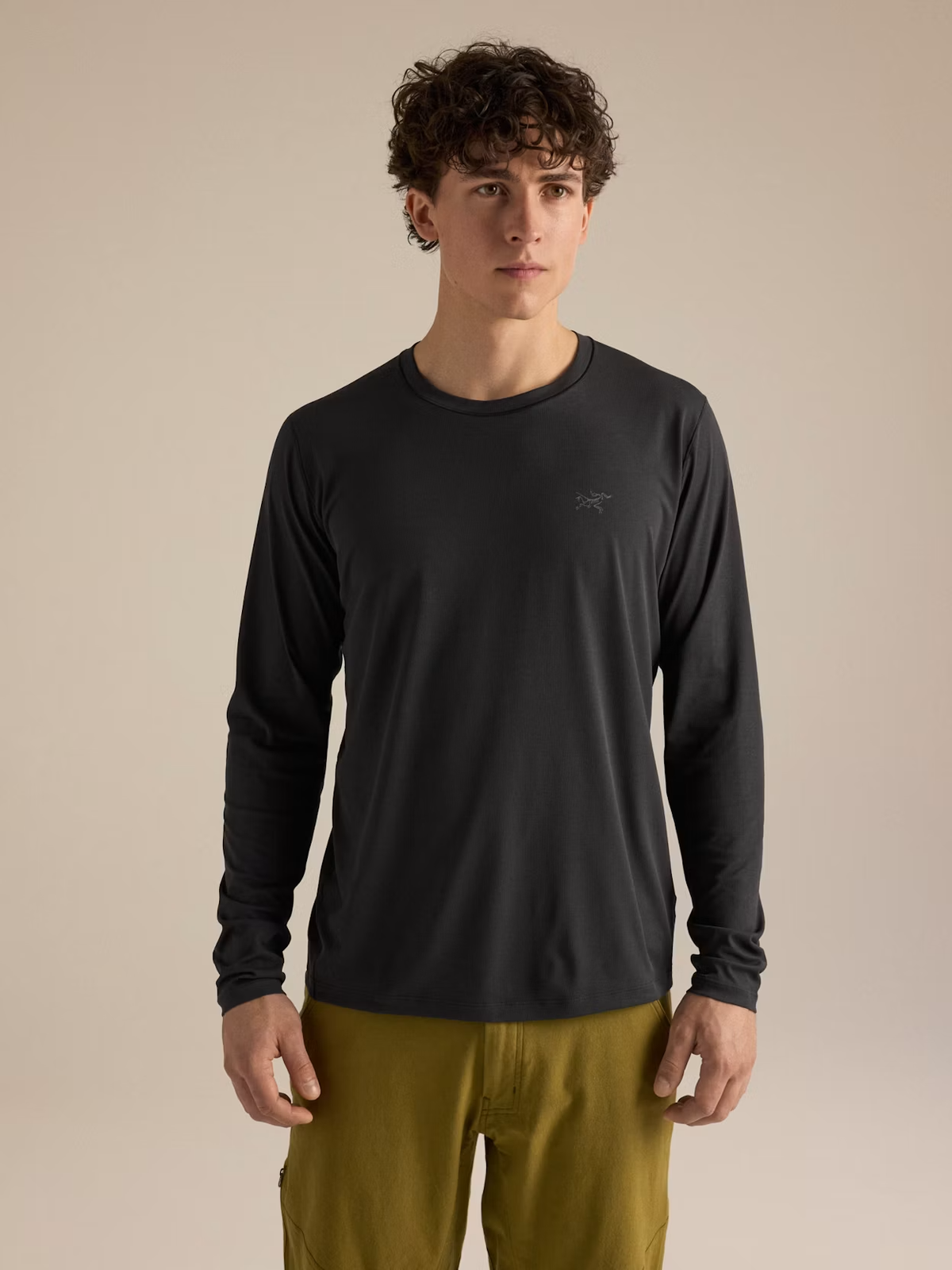 A man wearing a dark charcoal long-sleeve athletic shirt with a small Arc'teryx logo on the left chest, paired with olive green pants, standing against a solid beige background. - Arc'teryx Cormac Crew Neck Shirt Long Sleeve (Men's) - Black