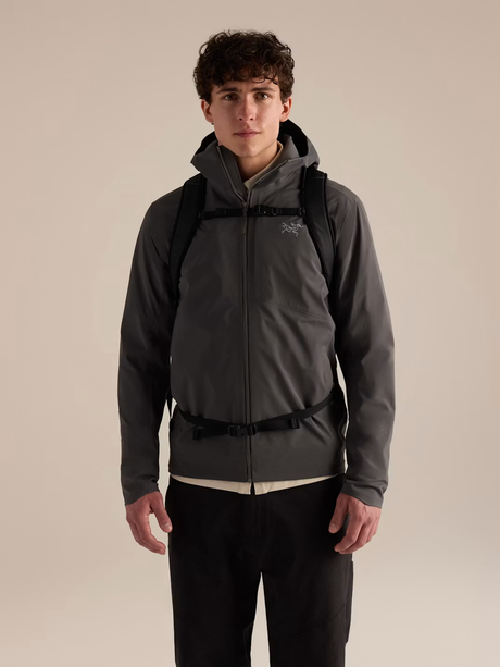 A man wearing a dark gray Arc'teryx hooded jacket with black straps and black pants, standing against a solid beige studio background. - Arc'teryx Mantis 26L Backpack - Black