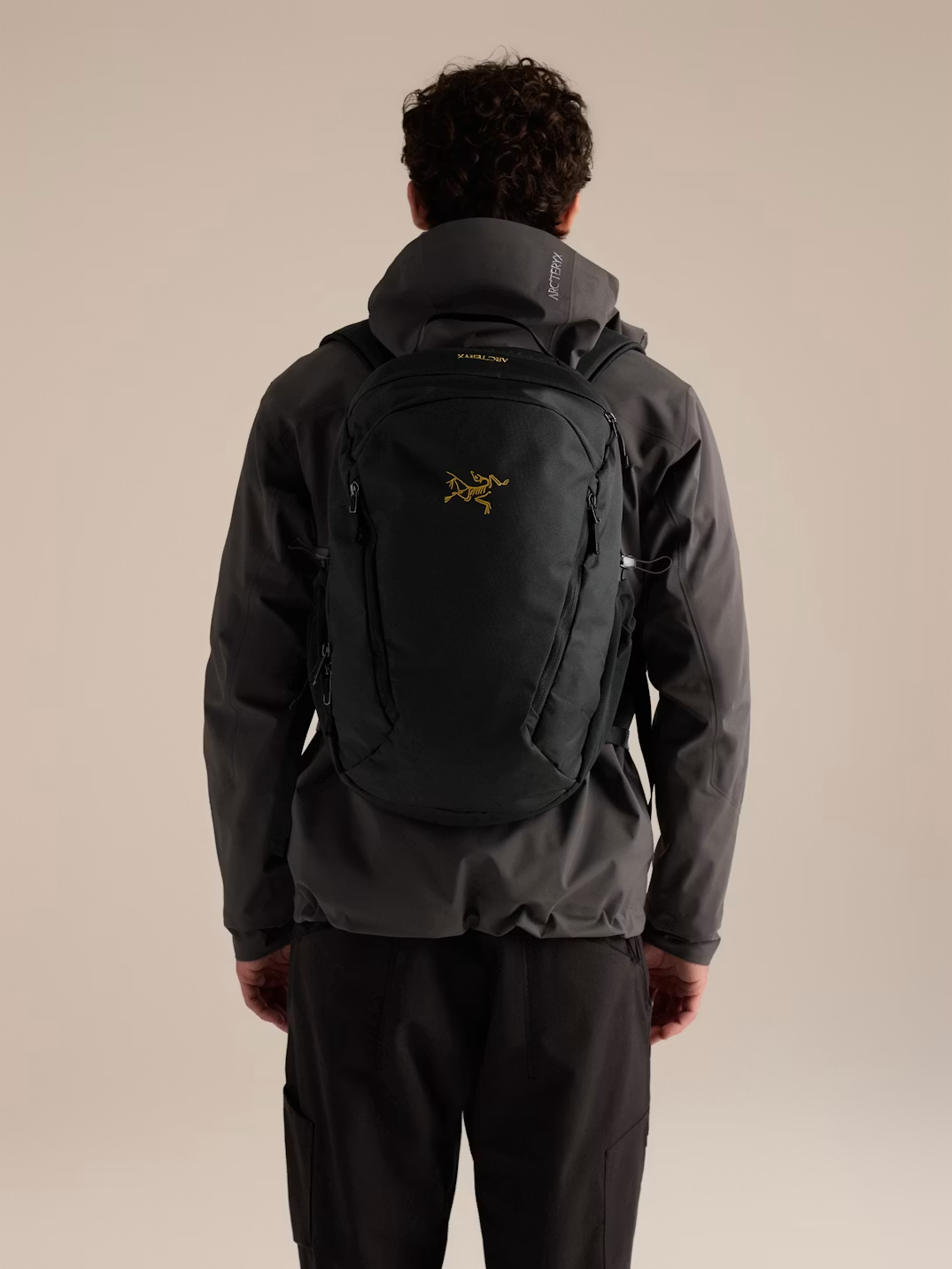 A man viewed from behind wearing a dark gray Arc'teryx jacket and black pants, carrying a black Arc'teryx backpack with a gold logo on the back. - Arc'teryx Mantis 26L Backpack - Black