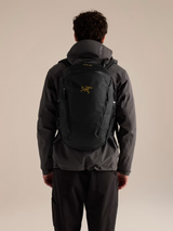 A man viewed from behind wearing a dark gray Arc'teryx jacket and black pants, carrying a black Arc'teryx backpack with a gold logo on the back. - Arc'teryx Mantis 26L Backpack - Black