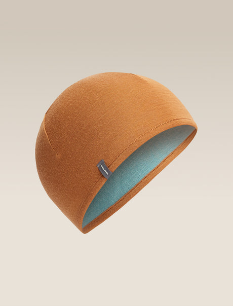 A warm camel-colored knit beanie hat with a light blue interior lining, shown floating against a neutral beige background. - Icebreaker Merino 200 Pocket Hat (Unisex) - Hydro/Trail