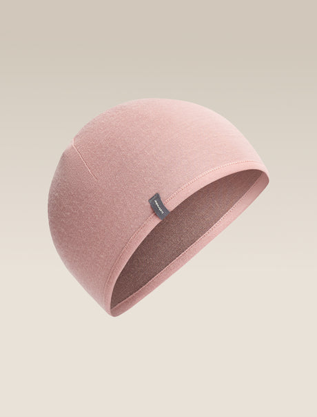 A dusty pink knit beanie hat with a small dark gray brand tag on the side, shown against a solid light beige background. - Icebreaker Merino 200 Pocket Hat (Unisex) - Summit/Blush
