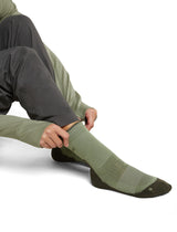 A person wearing an olive green sock with dark brown accents on the toe and heel, shown while pulling the sock up with both hands. The person is also wearing dark gray pants and an olive green long-sleeve top, set against a plain white studio background. - Icebreaker Merino Hike+ Light Crew Socks (Women's) - Lichen/Dark Loden
