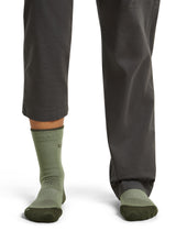 A close-up of a person's lower legs wearing dark gray pants and olive green crew socks with dark green toe and heel sections, shown against a plain white background. - Icebreaker Merino Hike+ Light Crew Socks (Women's) - Lichen/Dark Loden