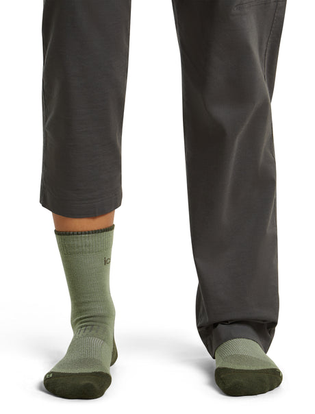 A close-up of a person's lower legs wearing dark gray pants and olive green crew socks with dark green toe and heel sections, shown against a plain white background. - Icebreaker Merino Hike+ Light Crew Socks (Women's) - Lichen/Dark Loden