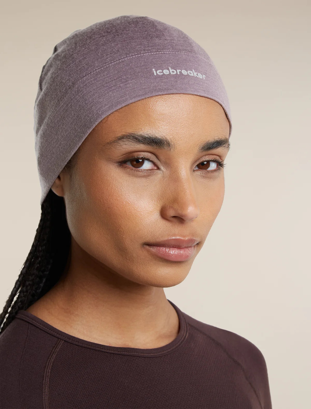 A close-up of a woman wearing a heathered mauve-purple athletic headband with the brand name 'icebreaker' printed in white on the front, paired with a dark brown athletic top against a solid beige background. - Icebreaker Merino 200 Oasis Beanie (Unisex) - Grey Plum