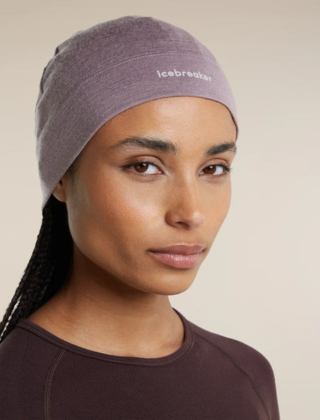 A close-up of a woman wearing a heathered mauve-purple athletic headband with the brand name 'icebreaker' printed in white on the front, paired with a dark brown athletic top against a solid beige background. - Icebreaker Merino 200 Oasis Beanie (Unisex) - Grey Plum