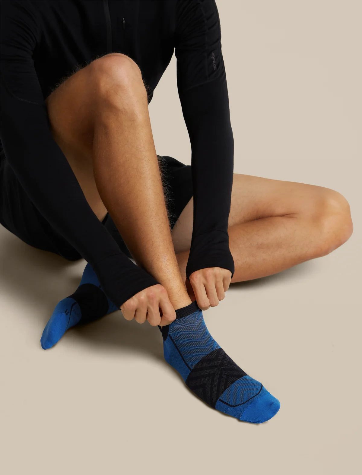A close-up of a person sitting and putting on a blue and black athletic crew sock, wearing a black long-sleeve top against a solid beige background. - Icebreaker Merino Run+ Ultralight Micro Socks (Men's) - Lazurite/Midnight Navy