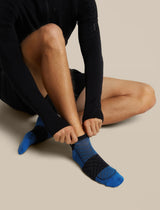 A close-up of a person sitting and putting on a blue and black athletic crew sock, wearing a black long-sleeve top against a solid beige background. - Icebreaker Merino Run+ Ultralight Micro Socks (Men's) - Lazurite/Midnight Navy