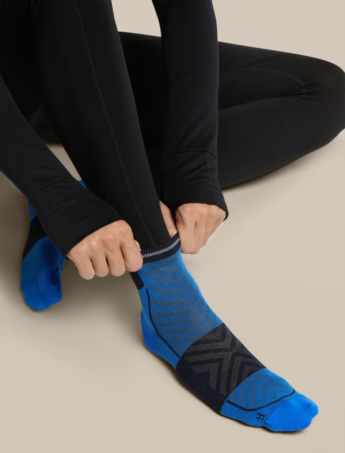 A close-up of a person wearing black leggings and adjusting a bright blue and black athletic sock on their foot against a plain beige background. - Icebreaker Merino Run+ Ultralight Mini Socks (Men's) - Lazurite/Midnight Navy