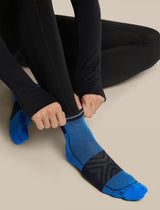 A close-up of a person wearing black leggings and adjusting a bright blue and black athletic sock on their foot against a plain beige background. - Icebreaker Merino Run+ Ultralight Mini Socks (Men's) - Lazurite/Midnight Navy
