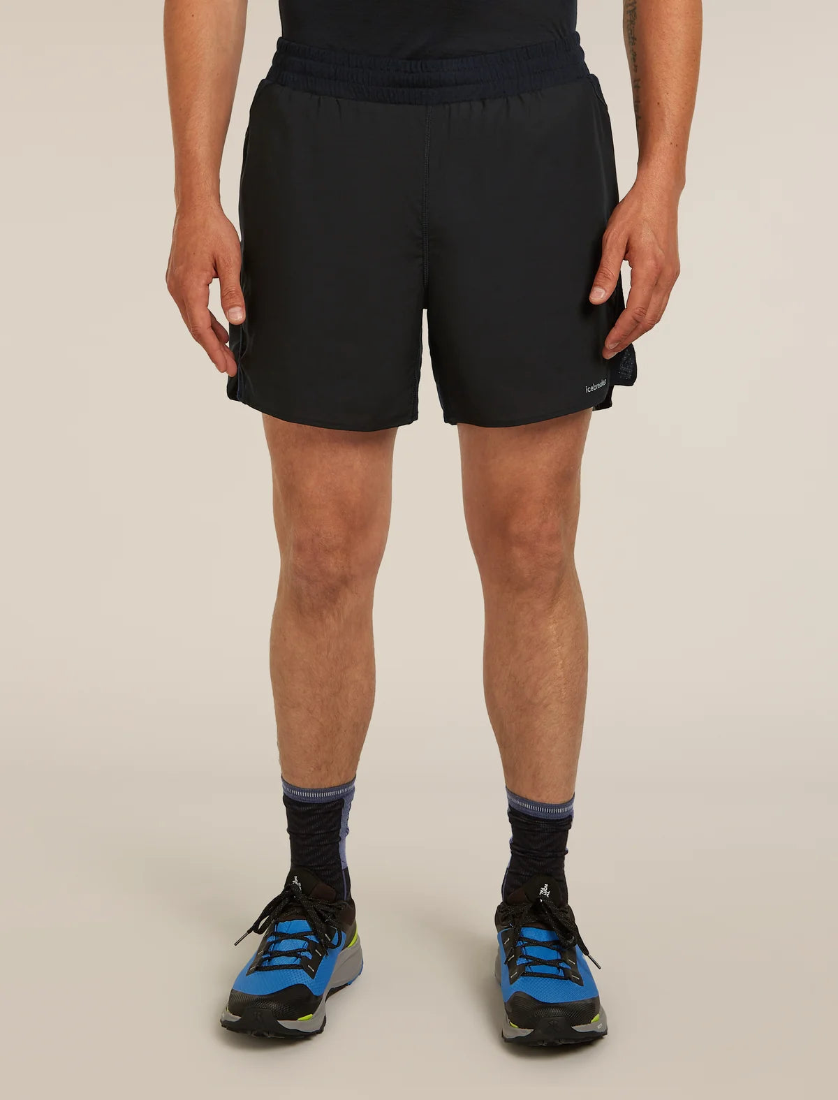 A person wearing black athletic shorts, black socks, and blue running shoes, shown from the waist down against a plain light beige studio background. - Icebreaker Merino Blend 125 ZoneKnit Speed 6" Shorts (Men's) - Black