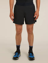 A person wearing black athletic shorts, black socks, and blue running shoes, shown from the waist down against a plain light beige studio background. - Icebreaker Merino Blend 125 ZoneKnit Speed 6" Shorts (Men's) - Black