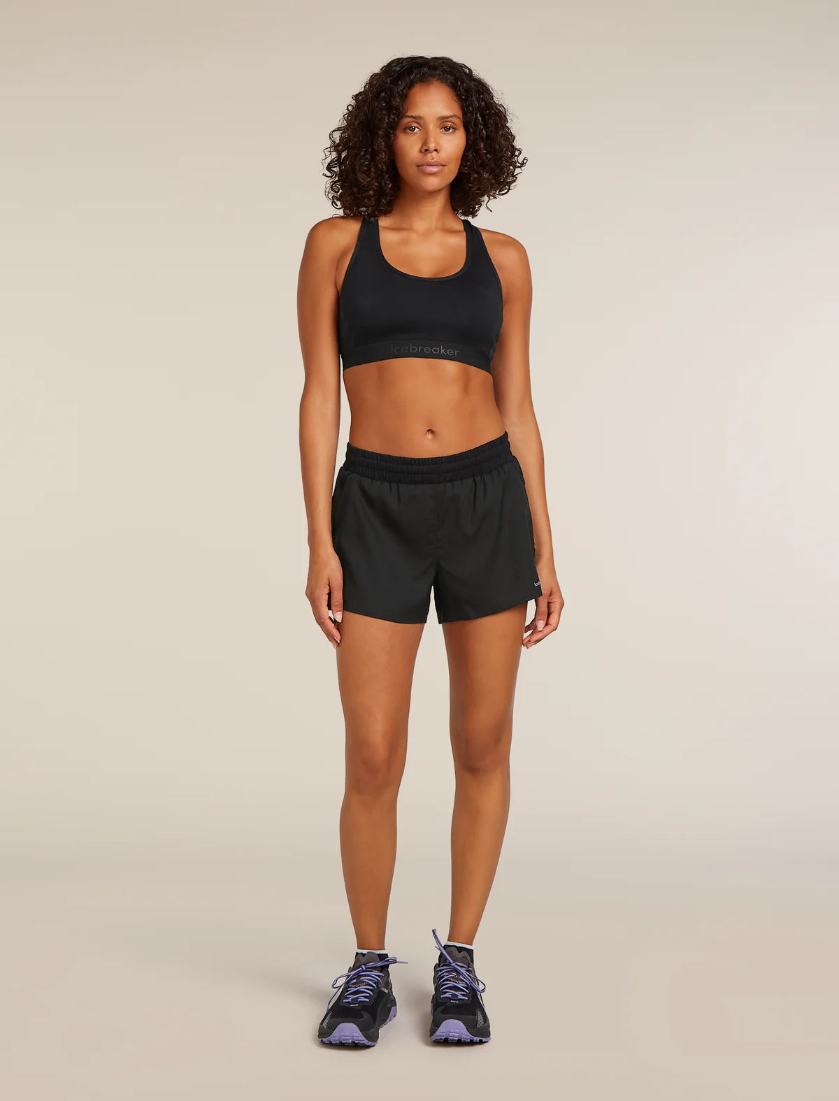 A woman stands against a plain beige studio background wearing a black sports bra and black athletic shorts, with dark running shoes. - Icebreaker Merino Blend 125 ZoneKnit Speed 3" Shorts (Women's) - Black