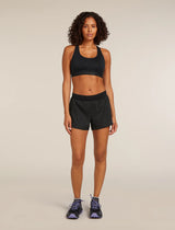 A woman stands against a plain beige studio background wearing a black sports bra and black athletic shorts, with dark running shoes. - Icebreaker Merino Blend 125 ZoneKnit Speed 3" Shorts (Women's) - Black