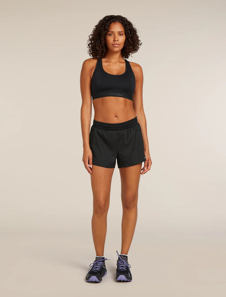 A woman stands against a plain beige studio background wearing a black sports bra and black athletic shorts, with dark running shoes. - Icebreaker Merino Blend 125 ZoneKnit Speed 3" Shorts (Women's) - Black