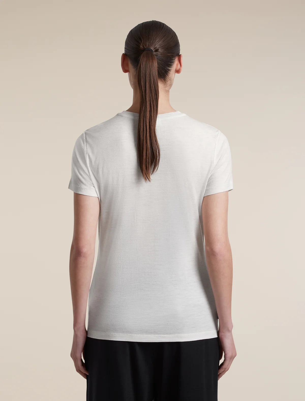 A woman viewed from the back wearing a plain white short-sleeve crew-neck T-shirt and black pants, standing against a light beige studio background. - Icebreaker Merino 150 Tech Lite Short Sleeve T-Shirt (Women's) - Ecru Heather