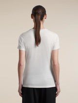 A woman viewed from the back wearing a plain white short-sleeve crew-neck T-shirt and black pants, standing against a light beige studio background. - Icebreaker Merino 150 Tech Lite Short Sleeve T-Shirt (Women's) - Ecru Heather